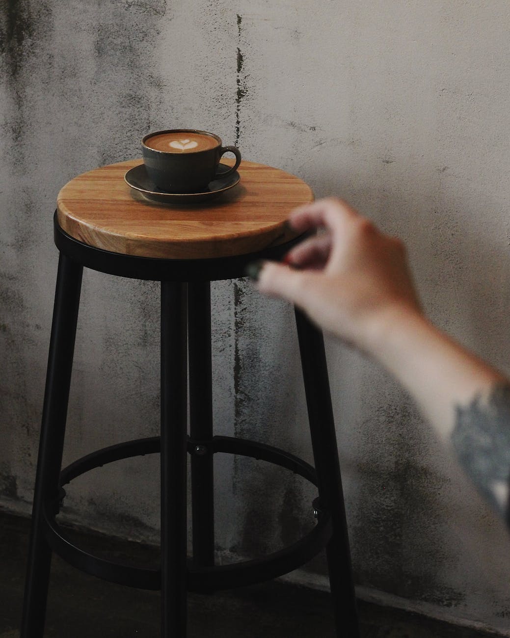 A craving hand reaching for a black ceramic coffee mug containing a latte and sitting on a stool.
